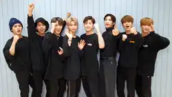 The eight members of Stray Kids, all in black, look forward and cheer