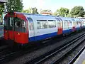 A side view of a tube train with red front. The sides of the carriages are white with a blue strip along the bottom quarter and red doors.