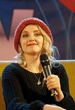 A caucasian female with light blonde hair, wearing a pink beanie hat and sitting on a couch while holding a microphone.
