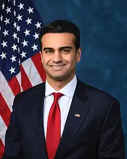 A headshot of Hamadeh smiling in front of an American flag