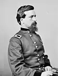 Profile of a dark-haired white man with a mustache and scraggly goatee, wearing a double-breasted military jacket and sitting with a military cap in his lap.