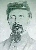Head of a white man with a goatee, wearing a flat cap and a high-collared jacket with a bowtie.