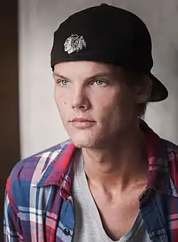 Avicii wearing a red flannel shirt over a gray t-shirt. He wears a black baseball cap, facing backwards. It bears the NHL's Chicago Blackhawks logo.