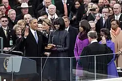 Barack Obama holds his right hand in the air as Chief Justice John Roberts administers the oath of office to him. Michelle Obama looks at him with Malia and Sasha Obama.
