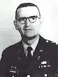 Head and shoulders of a middle-aged man with closely cropped hair and horn-rimmed glasses. He is wearing a dark military jacket with a parachute badge on the left breast and a Christian cross under a "U.S." pin on each lapel.
