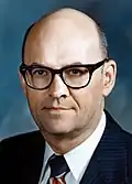 Color photo of a bald man wearing glasses and a suit with a striped tie