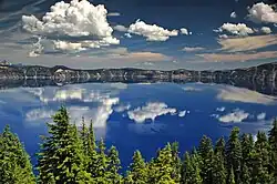Flat, blue lake clearly reflecting a cloudy sky, fronted by pine forest