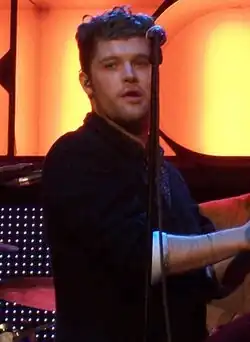 A thirty-six-year-old man is shown in three-quarter view. He is standing centre-stage, half-turned with hands clapped together. He is partly obscured by a microphone stand in front. Behind him is a drummer at his kit. Beyond that is the lighting, which includes the partly spelled letters R-O-C-K-C-O-R in large orange-yellow.