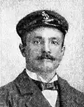 Head of a white man with handlebar mustache wearing a suit and bow tie and peaked cap.