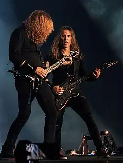 Dave Mustaine and Kiko Loureiro at a concert. They are both playing guitars.