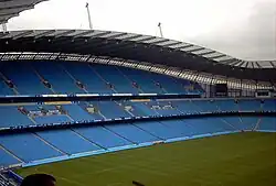 Roughly the same camera position shows grass up to the blue seats of the stands. The stand is now split into three levels of permanent seating.