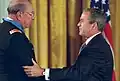 Profiles of two men shaking hands. The man on the right, wearing a dark suit, has his left hand on the other man's shoulder. The man on the left is taller and wearing glasses, a military jacket, and a light-blue ribbon around his neck.