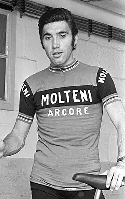 Merckx holding a bicycle. His shirt says "Molteni Arcore", and his hair is slicked back.