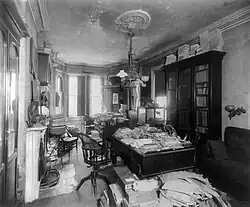 This picture is of a room with bookcases against the walls and two Louis-the-16th style desks in the middle of the room. Old mail is piled on the desks and on the floor by the desks.
