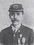 Head and shoulders of a young man with a mustache, wearing a military cap and a jacket with three medals pinned to the left breast.