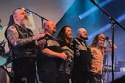 Five rock musicians side by side on stage after a concert, holding around each other while smiling to the audience