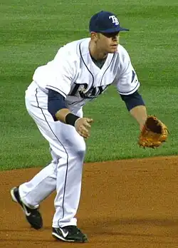 A man in a white and blue baseball uniform with the letters "TB" on his cap kneels, preparing to field a ground ball.