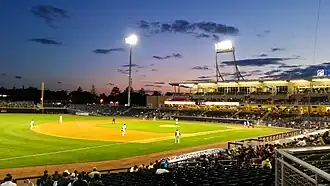 A view of the green baseball field from the third base side seats showing men in white baseball uniforms playing baseball as the sun has just set behind first base