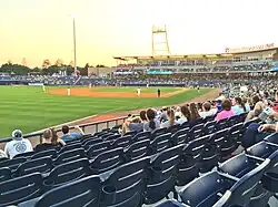 A view of a green baseball field from the third base side seats showing men in white baseball uniforms playing their positions with the late afternoon sun above the horizon