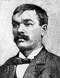 Head of a white man with mustache wearing a suit and bow tie.