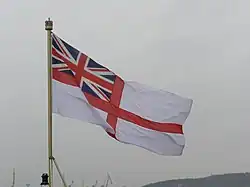 A flag waving in the wind. The flag is on a ship. The flag is white and has a red cross on it. There is a British flag in the top left of the flag.