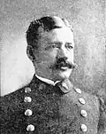 Head of a white man with neatly combed hair and a bushy mustache wearing a double-breasted military jacket with two columns of large buttons down the chest.