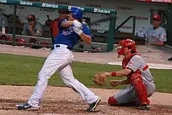 A man in a baseball uniform with white pants and a blue shirt swings while batting. The catcher in red and gray is also pictured.