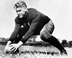 A uniformed but helmetless American Football player is shown on a football field. He is in a ready position, with legs in a wide stance and both hands on a football in front of him.