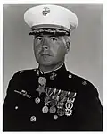 Head and shoulders of a middle-aged white man wearing a white peaked cap with oak leaf decorations on the visor and a dark military jacket with two rows of medals hanging from ribbons pinned to his chest and another medal hanging from his neck.