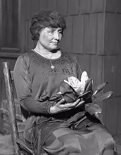 A woman with full dark hair and wearing a long dark dress, her face  in the partial profile, sits in a simple wooden chair. A locket hangs from a slender chain around her neck; in her hands is a magnolia, its large white flower surrounded by dark leaves.