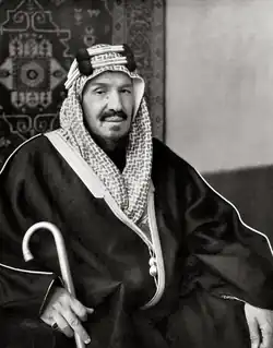 A black and white photograph of King Abdulaziz bin Abdul Rahman Al Saud is sat down, holding a cane and wearing traditional Arabian clothing. He is wearing a keffiyeh - a traditional Arab scarf - and an agal - a rope traditionally made from horse hair - on his head.
