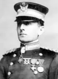 Head and shoulders of a white man standing erect, wearing a peaked cap over his eyes and a double-breasted military jacket with thick braided cords on the shoulders, three medals on the left breast, and a star-shaped medal hanging from his neck.