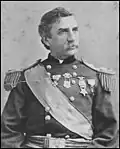 Head and torso of a white man with a mustache and wavy hair wearing an ornate military jacket. The jacket has fringed shoulder boards, a wide sash across the chest, and four medals pinned to the center and left half of the chest.