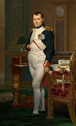 Full length portrait of Napoleon in his forties, in white and dark blue military dress uniform. He stands among rich 18th-century furniture. They have papers on them. He looks at the viewer. His hair is Brutus style, cropped close but with a short fringe in front. His right hand is in his waistcoat.