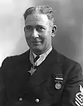 Head and shoulders of a white man with short bristly hair wearing a dark jacket with one round medal pinned to the left breast and a star-shaped medal hanging from a ribbon around his neck.