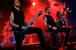 Three men are shown on an outdoor stage set during nighttime, the man on the left plays guitar, the man in the center plays bass guitar while the man on the far right yells into a microphone.