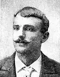 Head of a white man with neatly combed hair and handlebar mustache wearing a suit and tie.