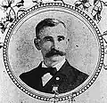 Head and shoulders of a white man with a wide bushy mustache, wearing a bow tie and dark suit with a medal pinned to the left breast. The portrait is surrounded by a circular frame and images of flowers.