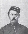 Head of a white man with a Van Dyke mustache and goatee, wearing a cap tilted on his head and a military jacket buttoned only at the neck.