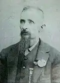 Head and shoulders of a white man with a long goatee, wearing a vest and suit coat with a flower pinned to the lapel.