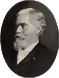 Profile of an older white man with a mustache and bushy goatee wearing a dark suit over a light-colored shirt and tie.