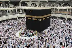 A large cubic monument covered in black silk (the Kaaba) is being circled by hundreds of Muslims dressed in white robes and shaven heads.
