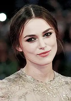 A portrait of Keira Knightley, wearing black tied-up hair, red lipstick, and a gold dress.