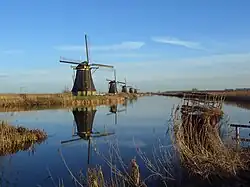 A windmill has large sails that use the force of the wind to turn heavy stone grinding surfaces, to grind grains into flour