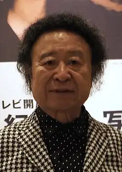 A Japanese man standing behind a poster-board, featuring a photograph of John Lennon and Yoko Ono kissing in New York.