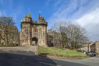 A pathway leading to a tall stone structure. There are two towers flanking a gateway, and walls continuing either side. In the foreground is a grassy area and trees.