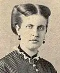 Photographic portrait of a young lady with dark-colored hair swept back and wearing a high-necked, Victorian era striped dress, dark earrings and a dark locket suspended around her neck on a ribbon