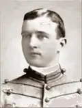 Head of a young white man with neatly combed hair wearing a jacket with lines of decorative piping running horizontally across the chest.