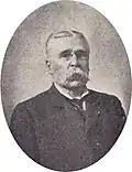 Head and shoulders of an older white man with a large drooping mustache wearing pince-nez glasses and a suit and tie. The portrait is surrounded by an oval frame and a depiction of an American flag.