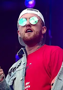 A young man on stage wearing round sunglasses and a brimmed cap turned backwards